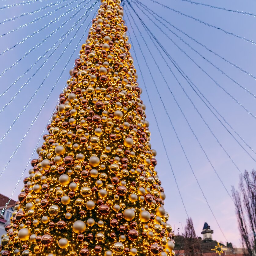 A large Christmas tree with golden ornaments on Mariahilferplatz in Graz, surrounded by a festive atmosphere and the clock tower in the background. | © Graz Tourismus - Mias Photoart