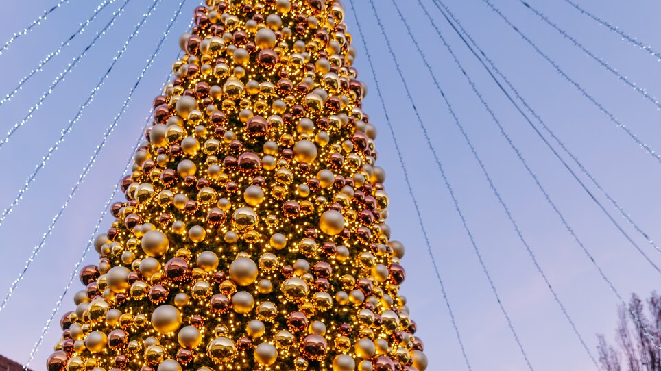 A large Christmas tree with golden ornaments on Mariahilferplatz in Graz, surrounded by a festive atmosphere and the clock tower in the background. | © Graz Tourismus - Mias Photoart