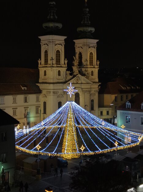 Beleuchteter Weihnachtsmarkt am Mariahilferplatz in Graz bei Nacht, umgeben von einer Kirche. | © Easy Conceptz GmbH