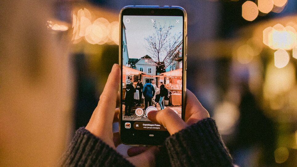 Eine Person fotografiert einen Weihnachtsmarkt mit Lichtern und Besuchern. | © Evgenia Rieger Photography