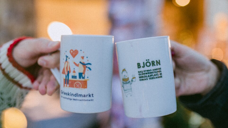 Zwei Personen stoßen mit Tassen am Grieskindlmarkt am Nikolaiplatz in Graz an. | © Evgenia Rieger Photography