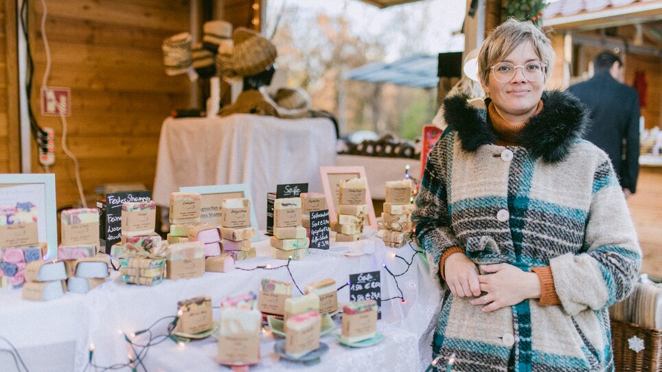 Eine Frau steht vor einem Marktstand mit handgemachten Seifen. | © Evgenia Rieger Photography