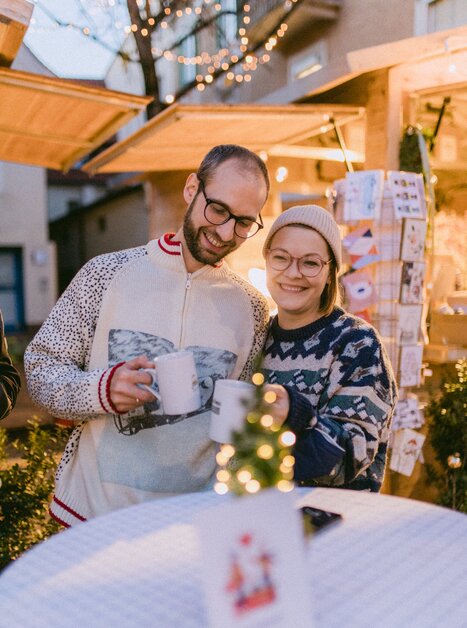 A couple with mugs in front of a Christmas market stand, festive lights in the background. | © Evgenia Rieger Photography