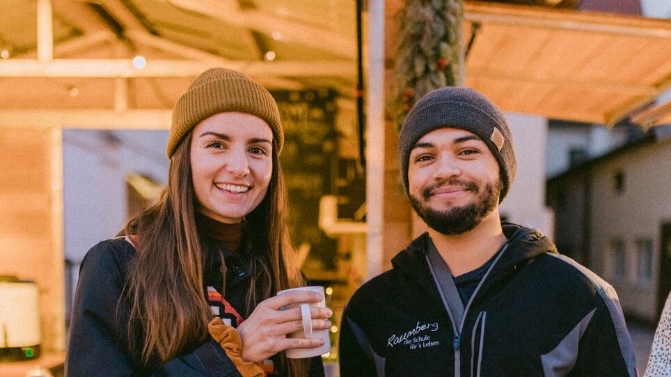 Ein Paar in Winterkleidung hält Tassen in einer festlich beleuchteten Umgebung. | © Evgenia Rieger Photography