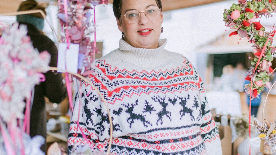 A person in a patterned sweater stands at a market stall surrounded by Christmas decorations and flowers. | © Evgenia Rieger Photography