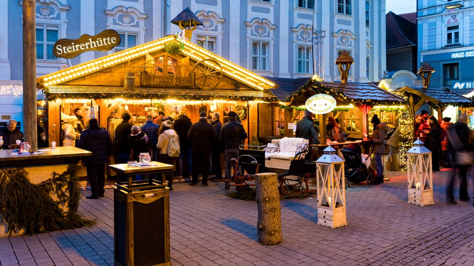 Evening atmosphere at the Christmas market with a festively lit wooden stall and crowds of people. | © Graz Tourismus - Harry Schiffer