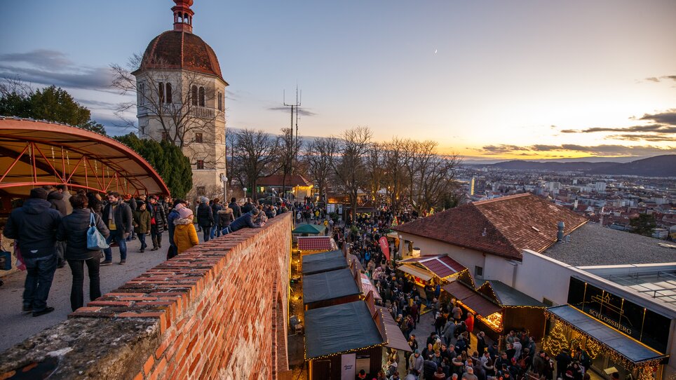 Blick auf den Weihnachtsmarkt am Schlossberg in Graz mit vielen Menschen und einem beeindruckenden Sonnenuntergang im Hintergrund. | © ivents - Erwin Scheriau