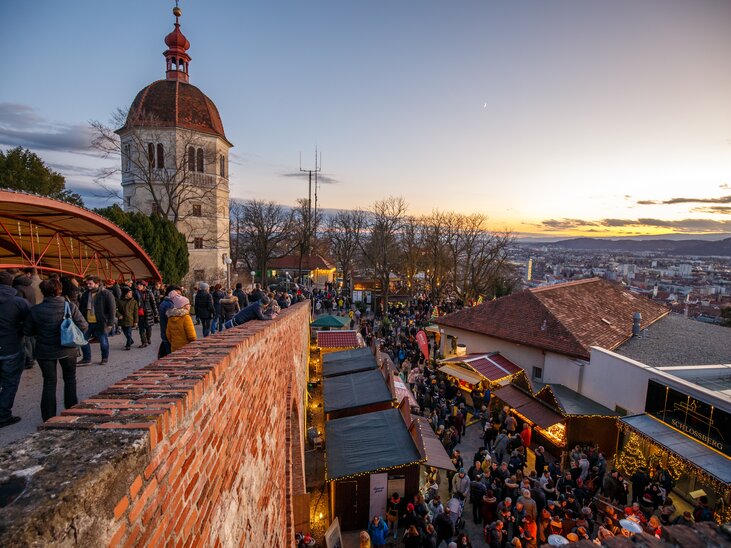 Blick auf den Weihnachtsmarkt am Schlossberg in Graz mit vielen Menschen und einem beeindruckenden Sonnenuntergang im Hintergrund. | © ivents - Erwin Scheriau