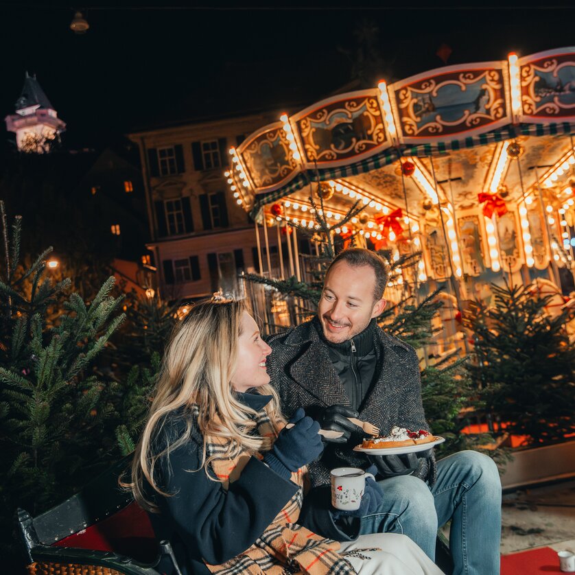 Ein Paar genießt Weihnachtsleckereien vor einem festlich beleuchteten Karussell in Graz. | © Mias Photoart