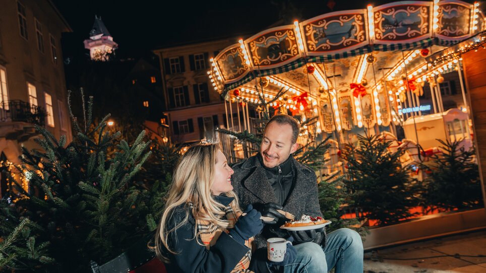 A couple enjoys Christmas treats in front of a beautifully lit carousel in Graz. | © Mias Photoart
