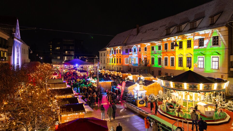 Illuminated Christmas market in Graz with festive stalls and carousel. | © Harry Schiffer