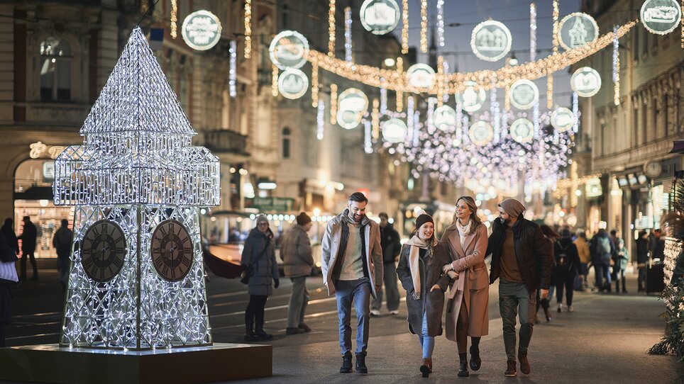 Beleuchteter Uhrturm im Vordergrund; vier Personen spazieren durch die festlich dekorierte Herrengasse in Graz. | © Robert Maybach