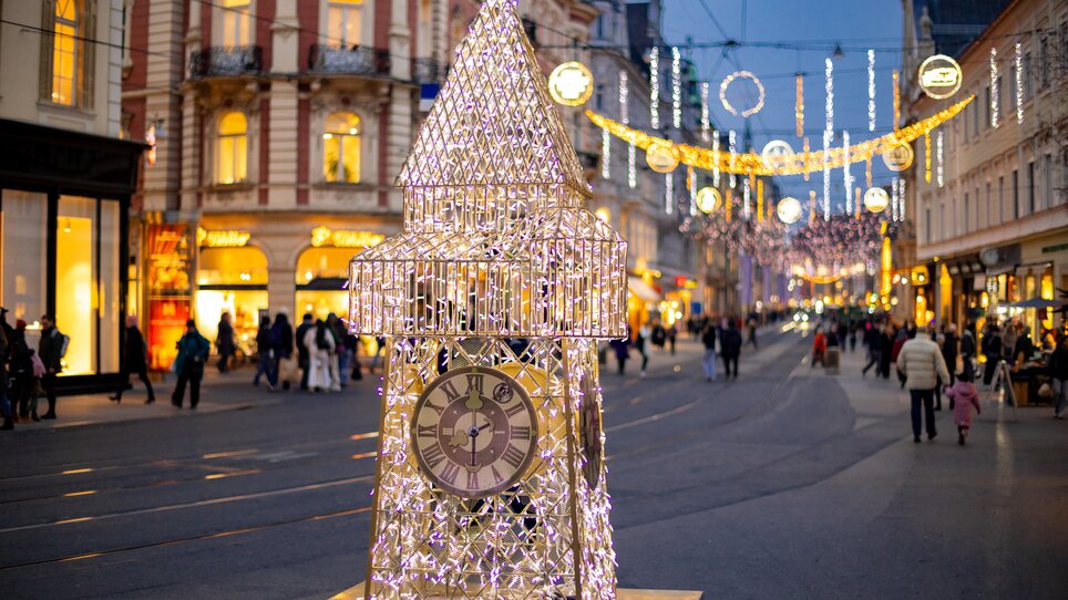 Ein beleuchteter Uhrturm in der Herrengasse in Graz mit Weihnachtsbeleuchtung zwischen den Häusern. | © Graz Tourismus - Harry Schiffer