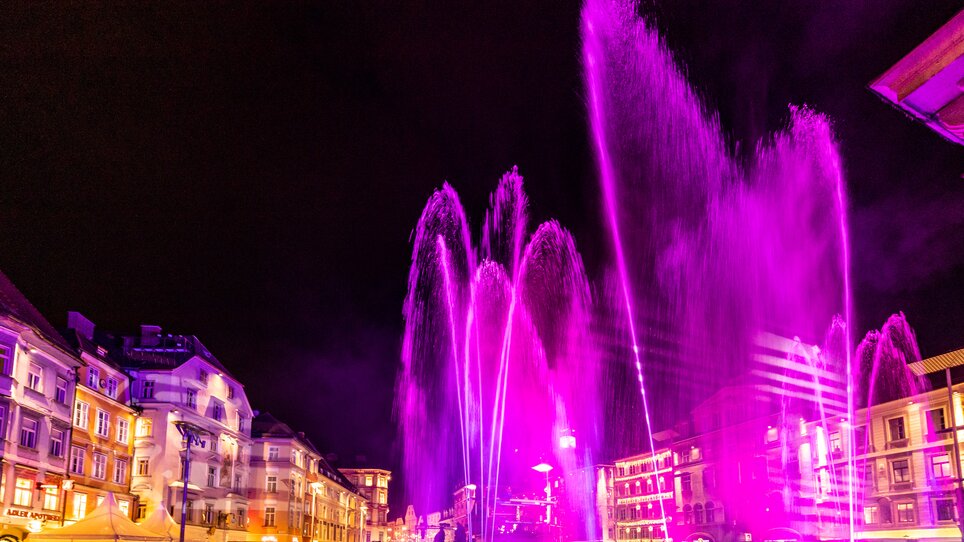 Fontane illuminate danzano in viola, circondate da edifici storici a Graz. | © Graz Tourismus - Harry Schiffer