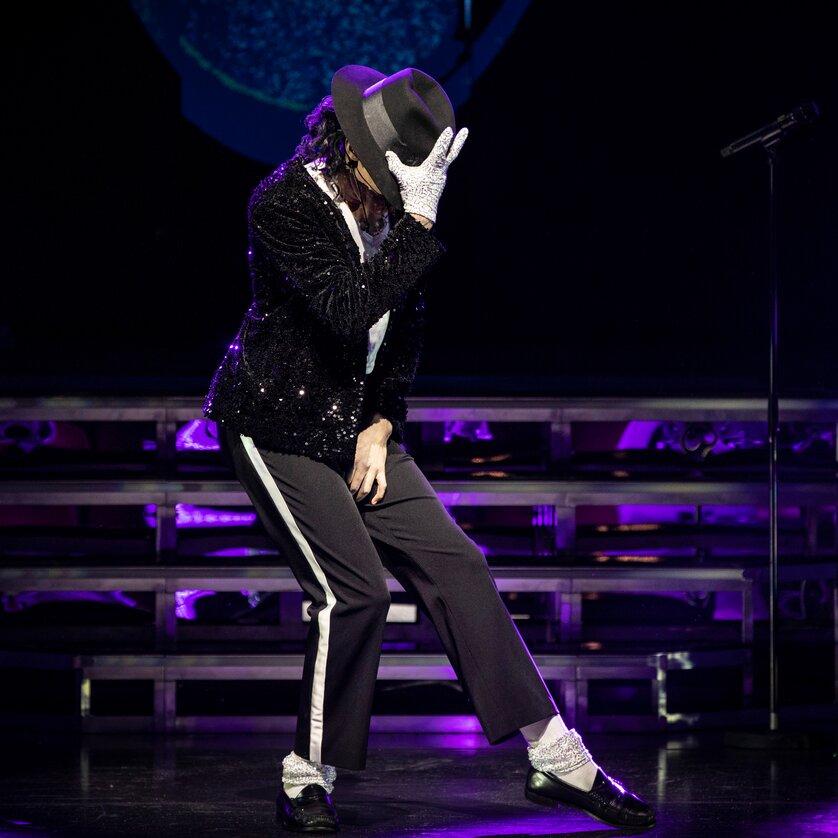 Un artista esegue una posa di danza sul palco, indossando un cappello e una giacca di paillettes.