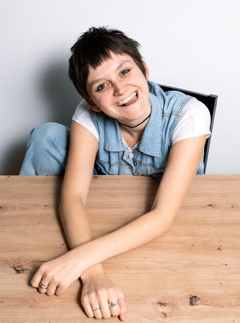 Giovane persona con capelli corti seduta a un tavolo, sorridente. | © Jasmin Pebal