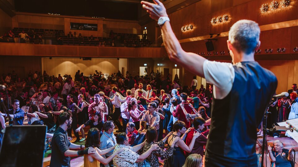 Ein Mann steht mit dem Rücken zur Kamera auf einer Bühne, im Zuschauerraum sieht man eine Polonaise aus Ballbesuchern | © Mario Sudy