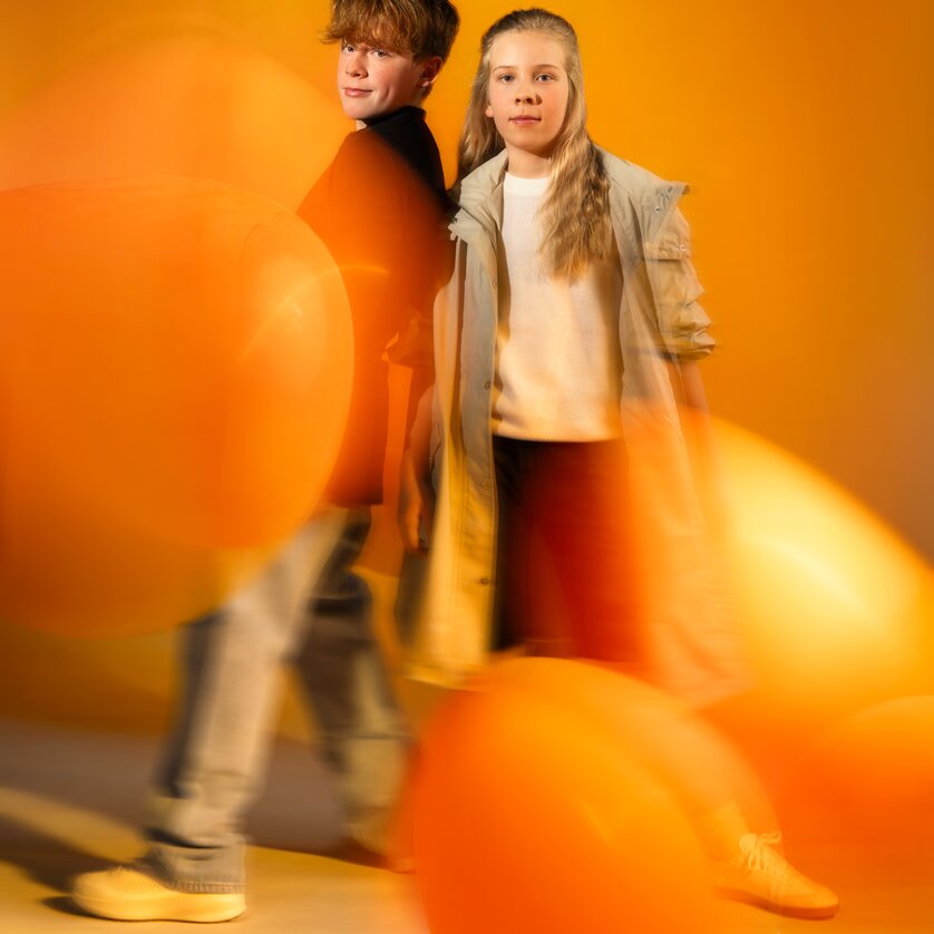 Zwei Kinder stehen mit bunten Luftballons vor einem orangefarbenen Hintergrund. | © Stella