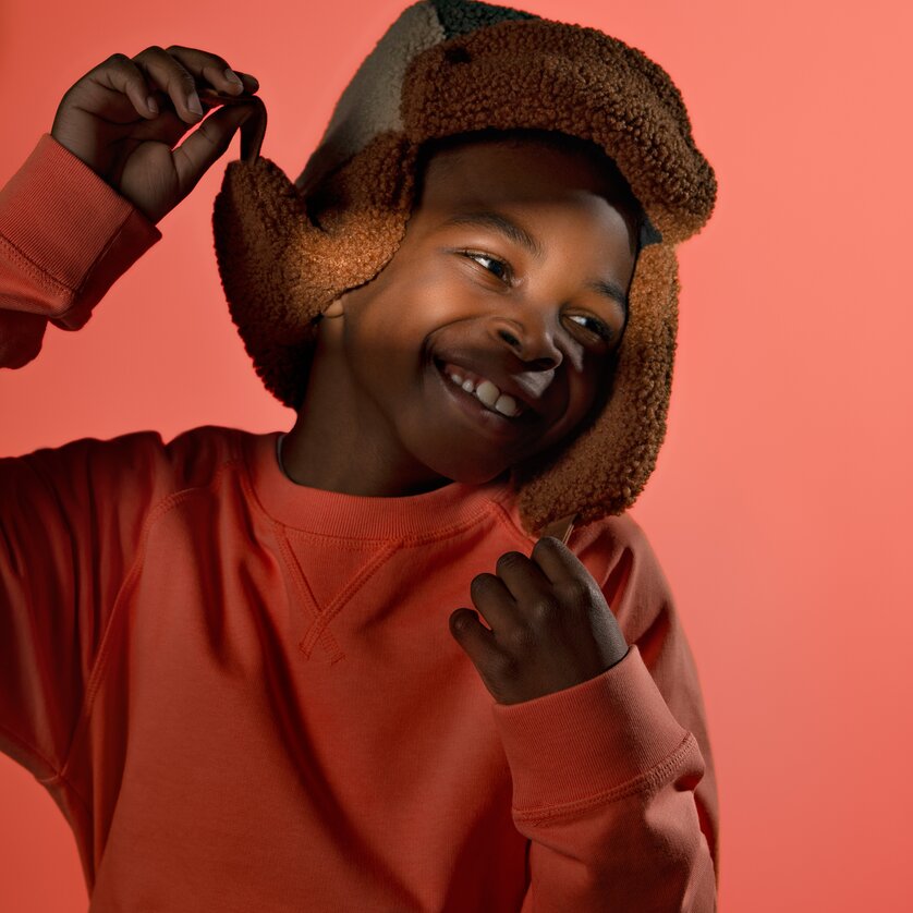 Ein fröhliches Kind in Orange trägt eine Plüschmütze und lächelt. | © Stella