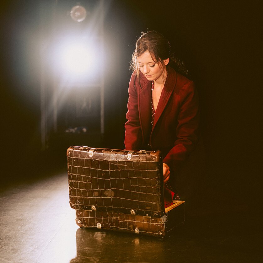 Eine Frau in einem roten Mantel kniet und öffnet einen Koffer auf einer Bühne, umgeben von Licht. | © Stella