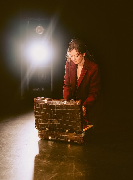 Eine Frau in einem roten Mantel kniet und öffnet einen Koffer auf einer Bühne, umgeben von Licht. | © Stella
