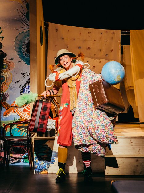 Ein fröhlicher Clown trägt viel Gepäck und einen Globus auf der Bühne. | © Stella