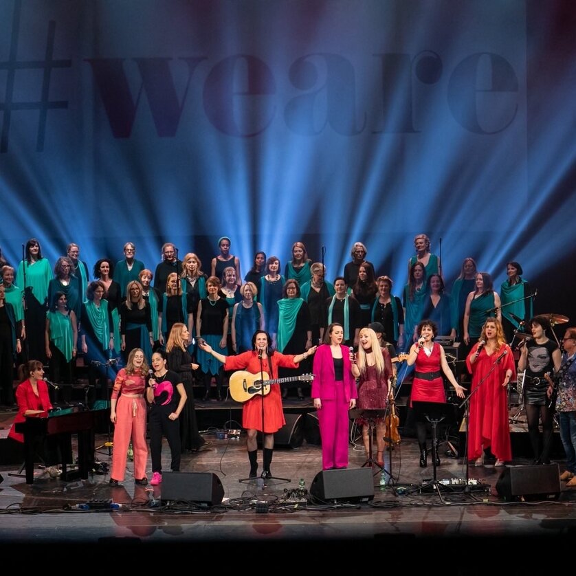 Große Gruppe von Sängerinnen und Musiker:innen bei Konzert mit Bühnenlicht und #weare-Backdrop | © Iva Lulic
