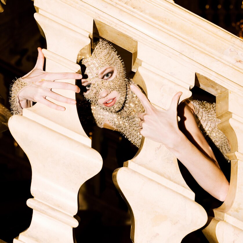 Eine Person mit einer goldenen Maske posiert hinter einer balustradenartigen Struktur. | © Oper Graz - Ingo Pertramer