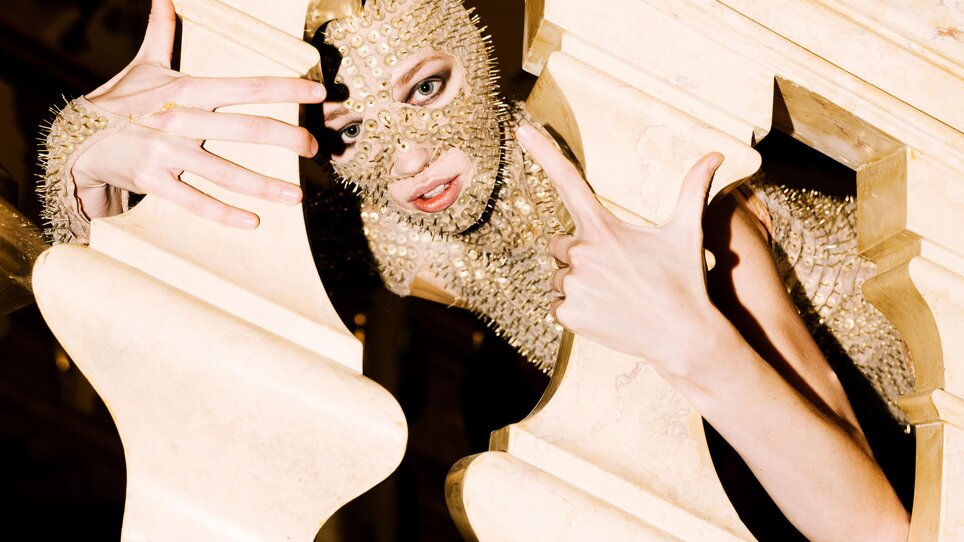 A person in a gold mask poses behind a balustrade-like structure. | © Oper Graz - Ingo Pertramer