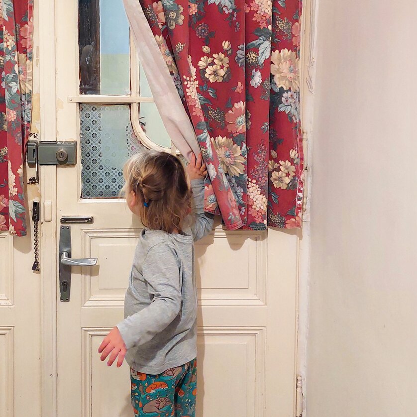 Ein Kind schaut durch die roten, blumengemusterten Vorhänge an einer Tür. | © Nina Strasser