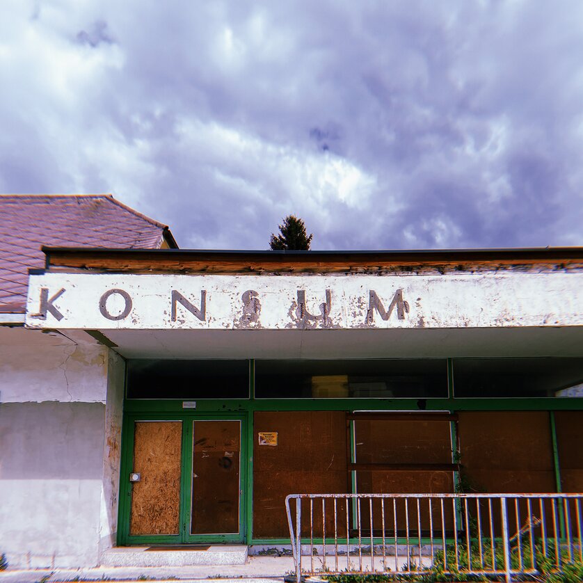 Eingang eines aufgegebenen Konsumgeschäfts mit beschädigtem Schild und Fenstern. | © Elmar Stengg