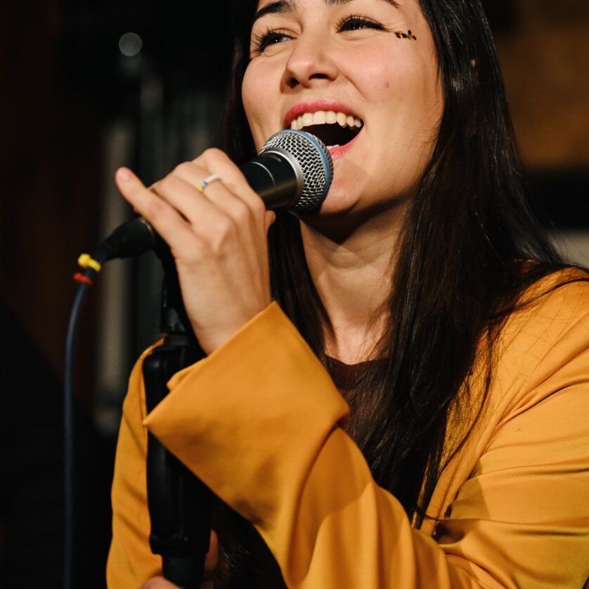 Eine Sängerin mit langen, dunklen Haaren singt ins Mikrofon. | © Bernhard Schaffer