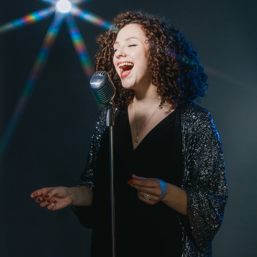 Donna con capelli ricci che canta in un microfono, effetti di luce sullo sfondo. | © Maria Frodl