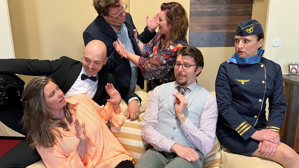 Sechs Schauspieler in einer chaotischen Szene, einige streiten, andere reagieren überrascht.