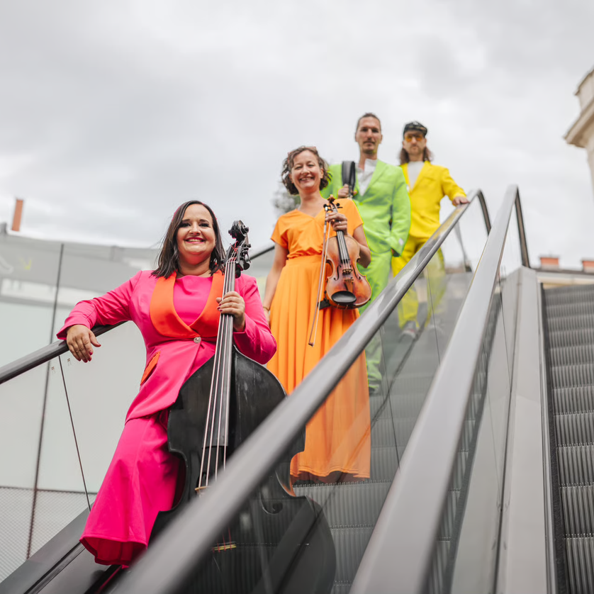 Four musicians in colorful outfits, one with a cello, posing on a staircase. | © Simon Reithofer