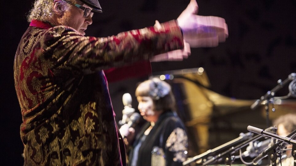 A musician conducts an orchestra while a singer performs in the background. | © Peter Purgar