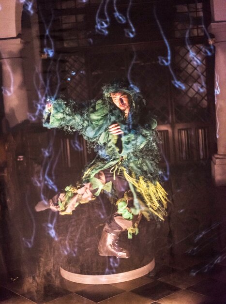Una persona in un costume verde fluttua sopra il suolo circondata da nebbia. | © Int. Bühnenwerkstatt