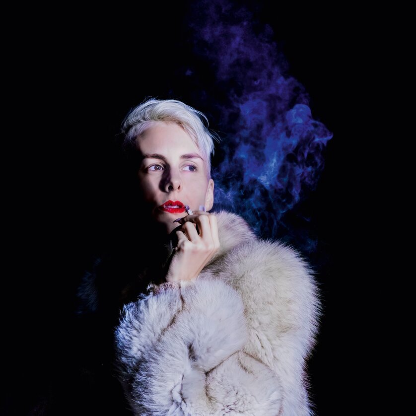 Ein Model mit blondem Haar in einem Pelzmantel posiert mit rauchigem Hintergrund. | © Jens Doerre