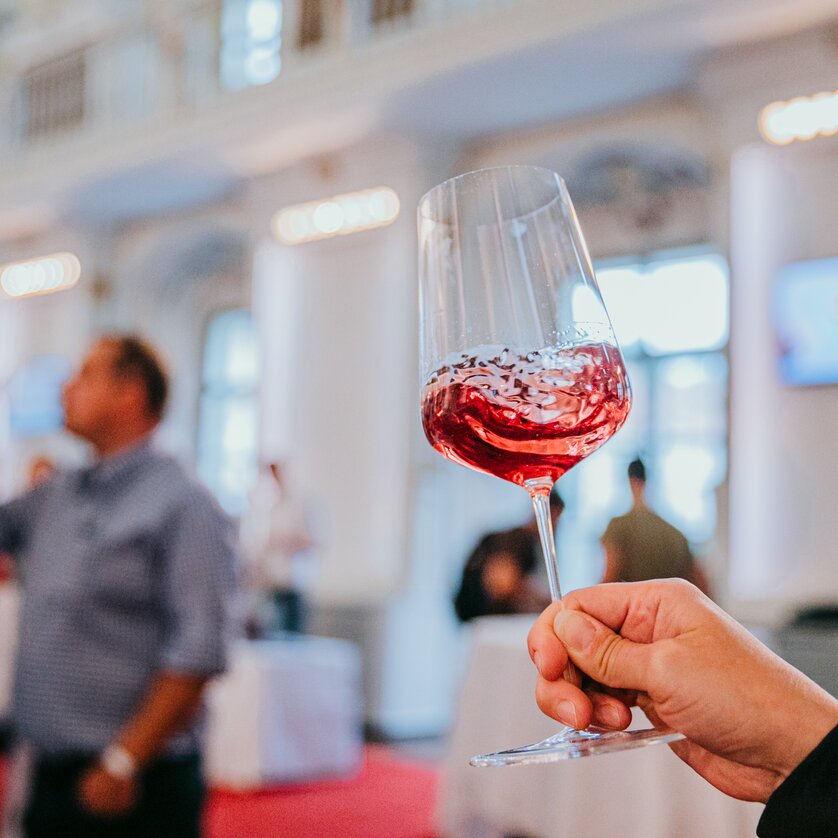A wine glass with shimmering red wine is held up to the camera. The wine swirls in the glass, people can be recognised blurred in the background. The atmosphere is festive and stylish. | © Johanna Lamprecht