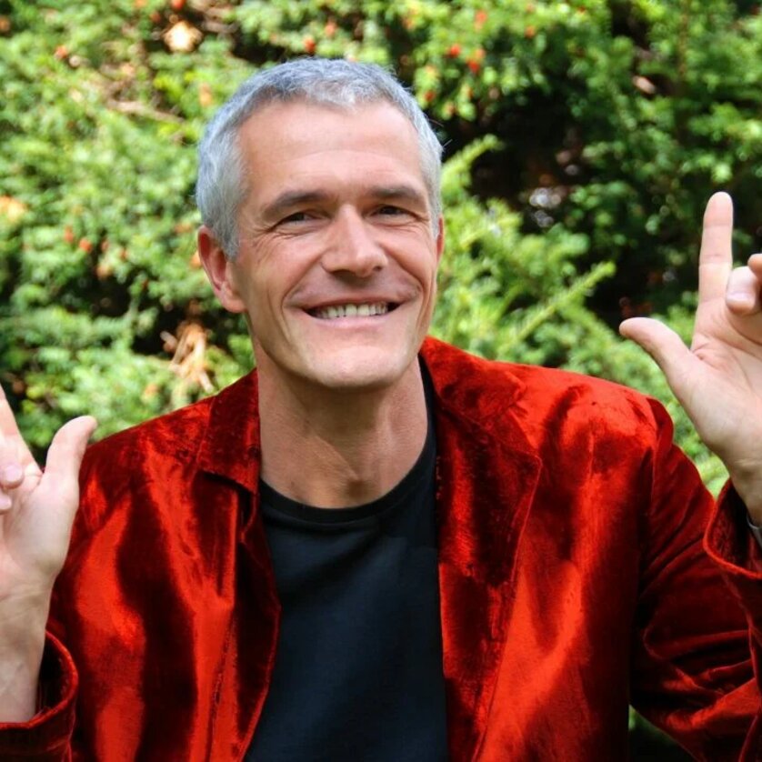 Ein Mann in einem roten Sakko lächelt und zeigt mit beiden Händen Zeichen. | © Beate Hofstadler