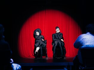 Zwei Performer in Masken auf der Bühne vor einem roten Vorhang. | © Daniel Kindler