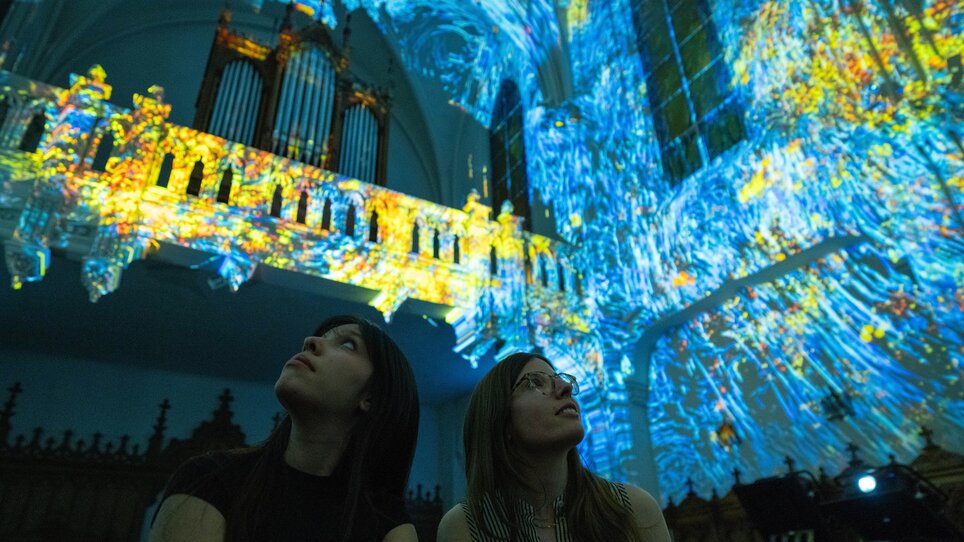 Zwei Personen bewundern eine farbenfrohe Lichtshow in einer Kirche. | © Projektil