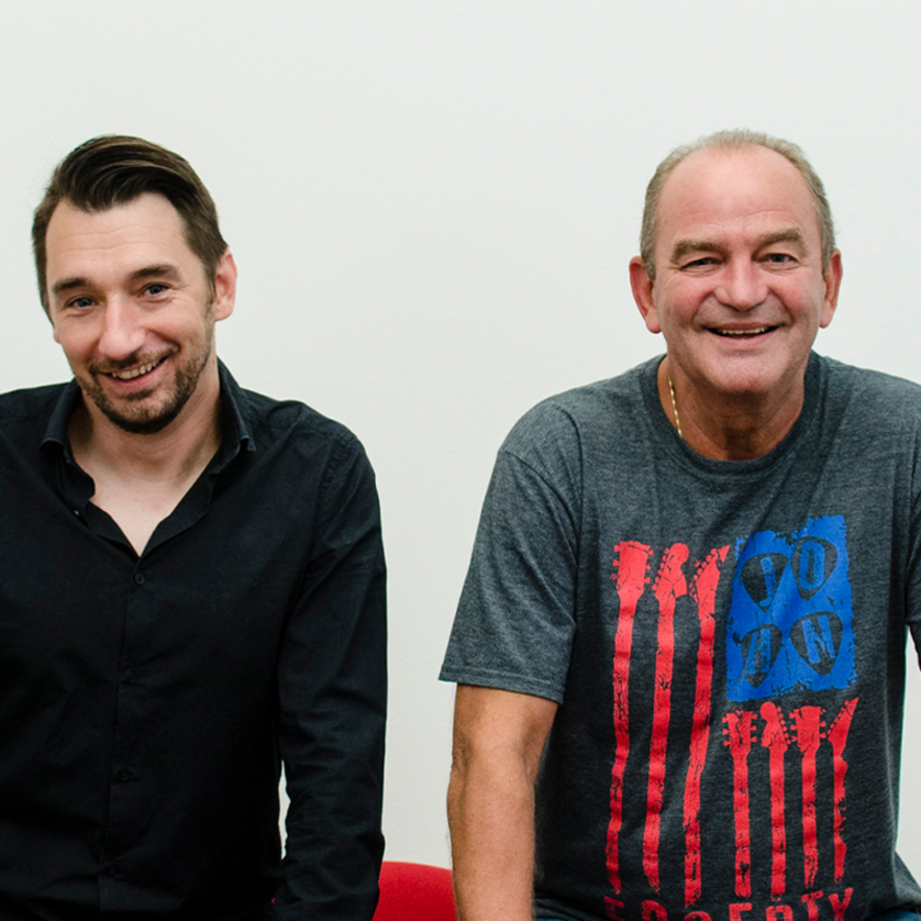 Zwei Männer stehen an einer Wand, einer trägt ein schwarzes Hemd, der andere ein graues T-Shirt mit roten und blauen Streifen. | © Bernhard Egger
