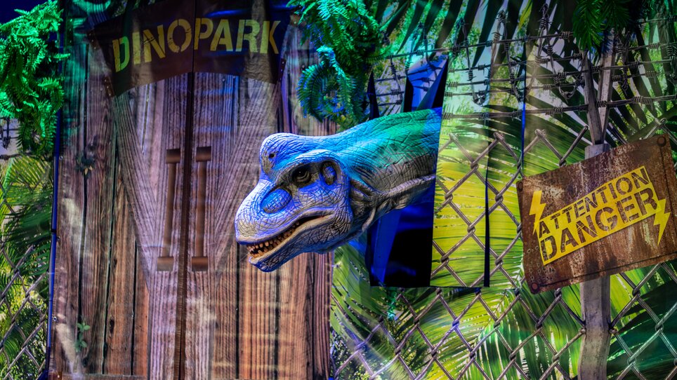 A dinosaur’s head peeks out from behind a backdrop at a dinosaur show. | © Timo Tutschek