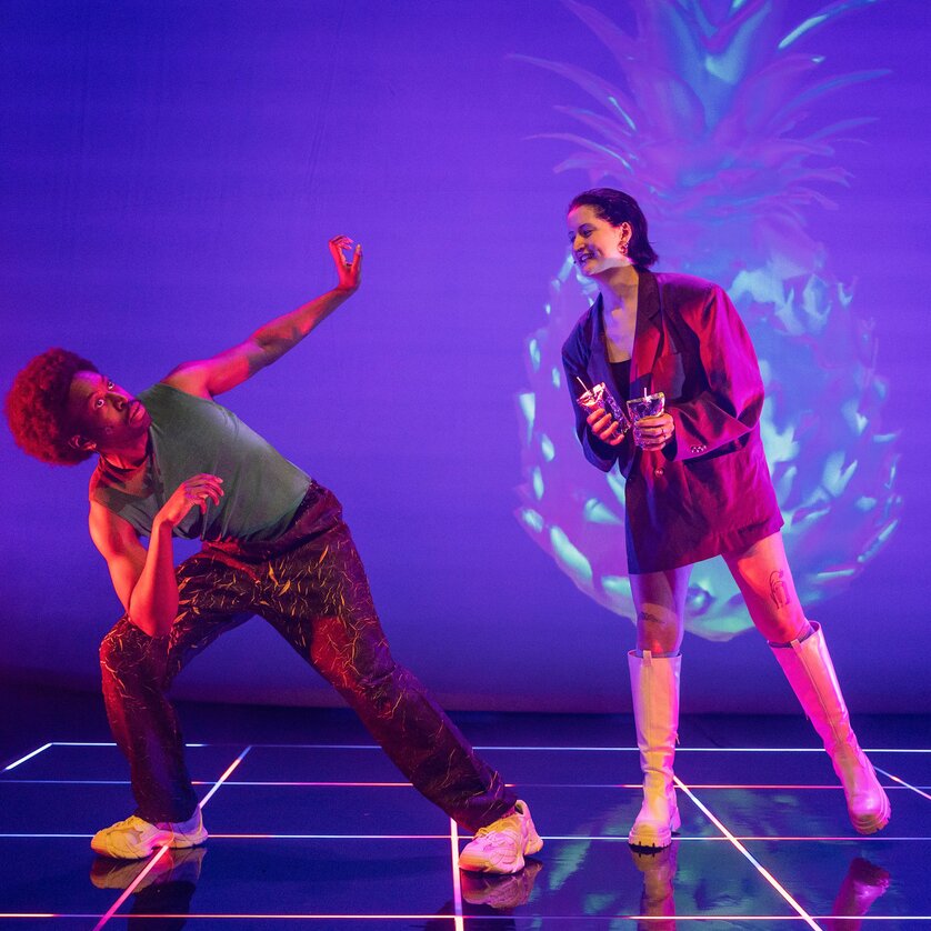 Zwei Darsteller interagieren auf einer Bühne mit farbenfrohen Lichtern. | © Phile Deprez