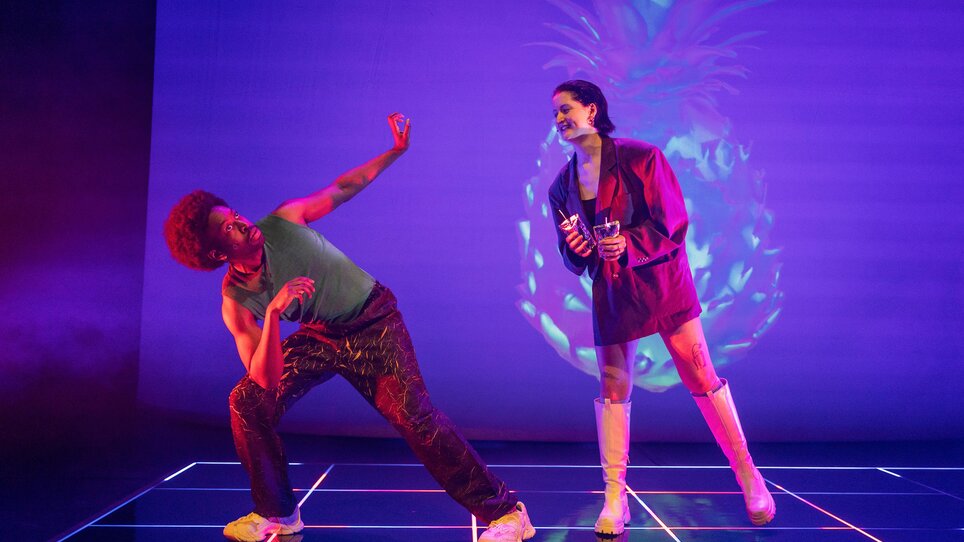 Zwei Darsteller interagieren auf einer Bühne mit farbenfrohen Lichtern. | © Phile Deprez