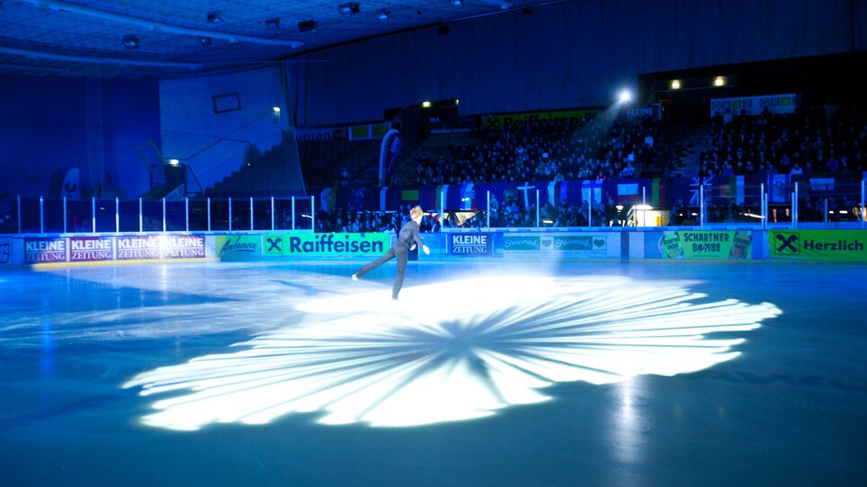 Eine Einzelkünstlerin führt eine elegante Eiskunstlauf-Performance auf einer beleuchteten Eisfläche vor Publikum. | © MCG - Martin Wiesner
