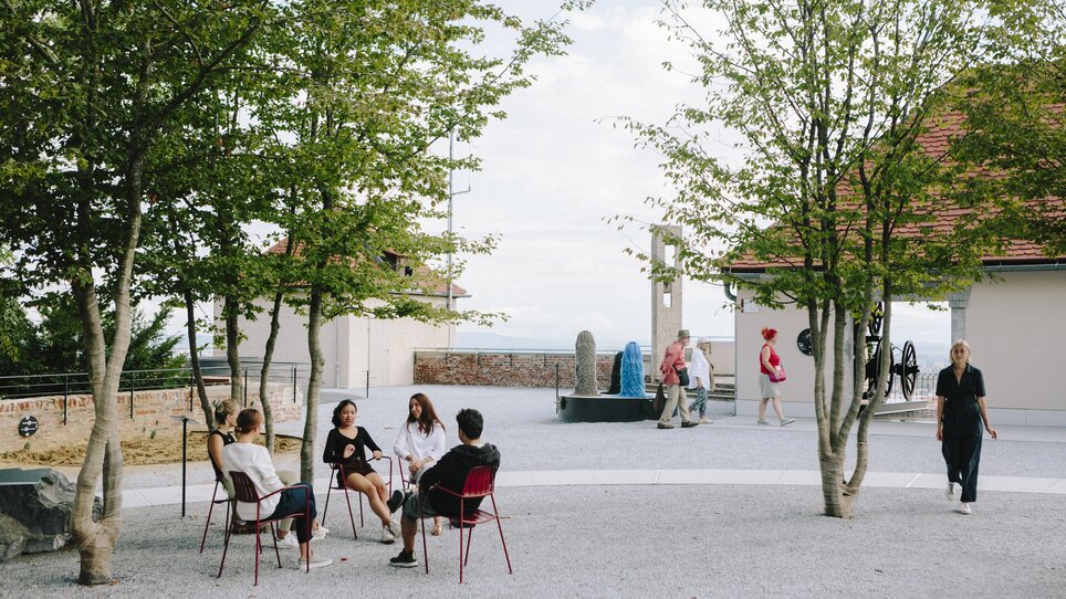 People in an outdoor area discussing, surrounded by trees and modern architecture. | © Sebastian Reiser