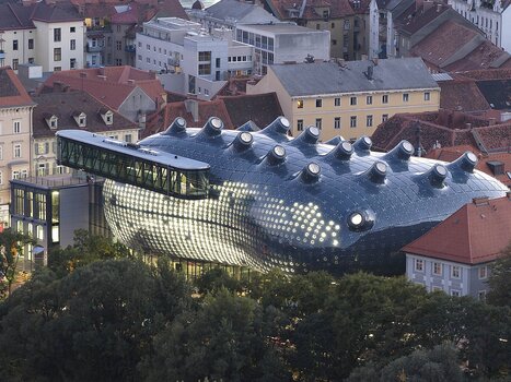 Futuristic, dark blue building with a glossy, bubble-like surface and multiple rounded skylights on the roof, surrounded by traditional red-roofed houses and trees; striking contrast between modern architecture and historic cityscape. | © UMJ -N. Lackner