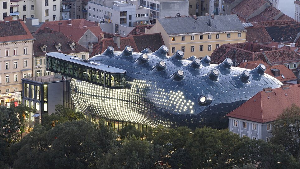 Futuristic, dark blue building with a glossy, bubble-like surface and multiple rounded skylights on the roof, surrounded by traditional red-roofed houses and trees; striking contrast between modern architecture and historic cityscape. | © UMJ -N. Lackner